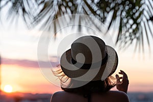 Back view of a beautiful girl in a straw hat against the background of the sea in branches of palm trees. Sunset beach. Summer