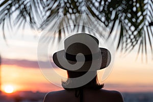 Back view of a beautiful girl in a straw hat against the background of the sea in branches of palm trees. Sunset beach. Summer