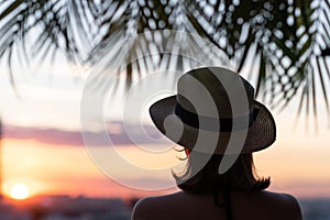 Back view of a beautiful girl in a straw hat against the background of the sea in branches of palm trees. Sunset beach. Summer