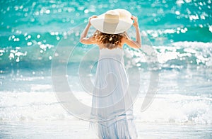 Back view beautiful girl with straw hat enjoying sunbath at beach. Young tanned woman enjoying breeze at seaside