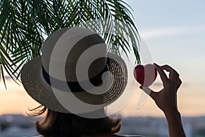 Back view of a beautiful girl in a straw hat holding red heart against the background of the sea in branches of palm trees. Sunset