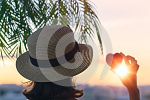 Back view of a beautiful girl in a straw hat holding red heart against the background of the sea in branches of palm trees. Sunset