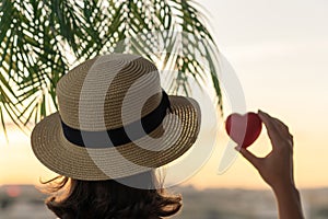 Back view of a beautiful girl in a straw hat holding red heart against the background of the sea in branches of palm trees. Sunset