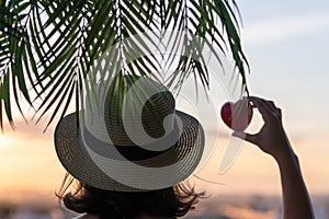 Back view of a beautiful girl in a straw hat holding red heart against the background of the sea in branches of palm trees. Sunset