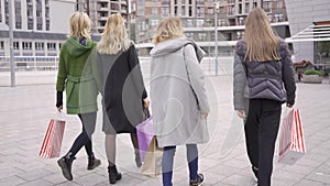 Back view beautiful girlfriends walking on the street talking happily. Four fashion women with shopping bags walking in