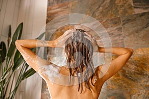 Back view of beautiful naked young woman taking shower in bathroom with marble tiles