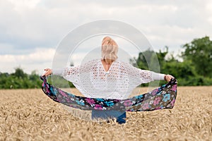 Back view of beautiful woman stand with spread arms, hold silk scarf, in golden wheat field with cloudy blue sky background