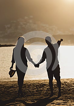 Back view of a beautiful young couple of teenagers holding their hands standing on the beach during sunset