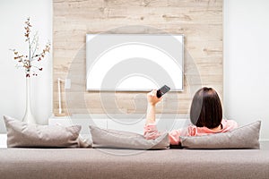Back view of beautiful young woman resting in apartment, sitting on sofa , watching TV, changing channel. Blank screen