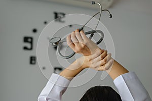 Behind young female doctor holding stethoscope standing and hand up for relaxing after finish work with clock on the wall.