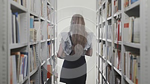 Back view of blonde student woman walking between the bookshelves in university library with many books in hands