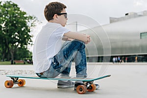 Back view. Boy in white T-shirt and blue jeans, sits on city street on skateboard. Space for text, logo, image. Mock up.
