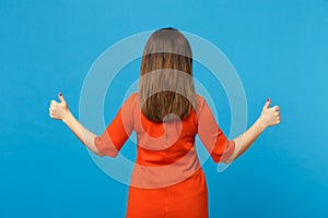 Back view brunette young woman wearing red orange dressshowing thumb up standing posing isolated over trendy blue wall