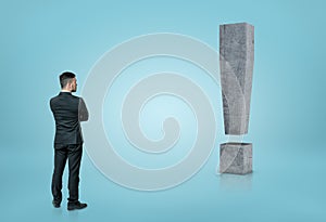 Back view of a businessman looking at big 3D concrete exclamation mark isolated on blue background