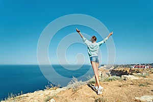 Back view of carefree young woman standing with raised hands on cliff edge with beautiful sea view and enjoying