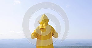 Back view of caucasian female hiker in yellow raincoat stands on top of a mountain