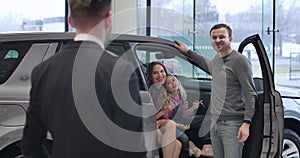 Back view of Caucasian male trader standing in car dealership and looking at happy young family in new automobile