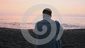 Back view of Caucasian man sitting on the beach by the sea alone at sunset. Sad male with depression, stress, melancholy