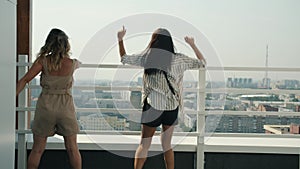 Back view of cheerful girls dancing at rooftop party doing high-five laughing enjoying view of metropolis