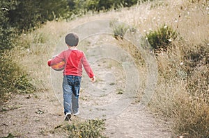 Back view of a child with a ball in his arm walking down a road