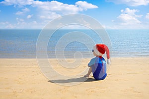 back view of child boy in santa hat, sunglasses and swimsuit sitting on the sand beach