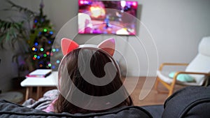 Back view of a child girl sitting on the couch and playing video game or watching cartoons on tv. Wireless funny pink