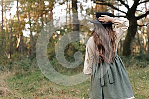 Back view of child girl in stylish canvas vintage style dress holds hand on black hat on nature background. Little model
