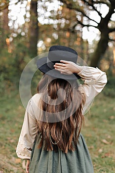 Back view of child girl in stylish canvas vintage style dress holds hand on black hat on nature background. Little model