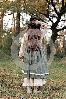 Back view of child girl in stylish canvas vintage style dress holds hand on black hat on nature background. Little model