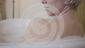 Back view close-up of Caucasian blond woman applying bath foam on shoulder in bathtub. Happy smiling lady relaxing in