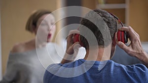 Back view close-up man putting on headphones ignoring woman yelling at background gesturing. Tired Caucasian husband