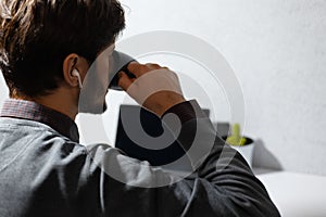 Back view; close-up of young man drinking coffee, using wireless earphones.