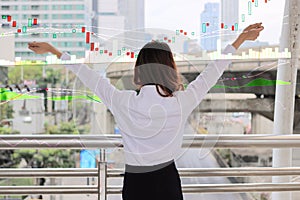 Back view of confident Asian business woman raising hand and looking far away in urban building city against growth stock chart gr