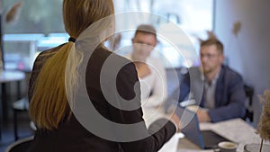 Back view of confident enthusiastic businesswoman talking to blurred men sitting at the table at the background