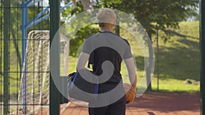 Back view of confident sportsman entering outdoor court with sports bag and ball. Young Caucasian basketball player