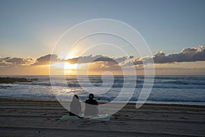 Back view of a couple sitting on the sandy beach watching the sea during cloudy sunset