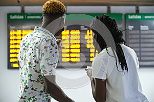 Back view of couple in a train station wearing masks, looking at their smartphone and the travel board