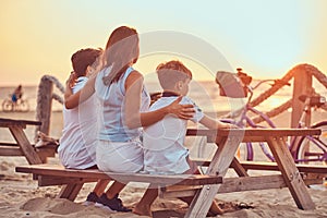 Back view of a cute family - mother with her sons sitting on a bench and looks at the bright sunset on the seacoast.