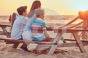 Back view of a cute family - mother with her sons sitting on a bench and looks at the bright sunset on the seacoast.