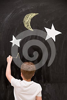 Back view on cute kid boy drawing stars with chalk on a black board with white stars and moon. Child writing on wall