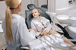 Back view of cute little girl sitting on dental chair and complaining about pain visiting pediatric dentist. Prevention