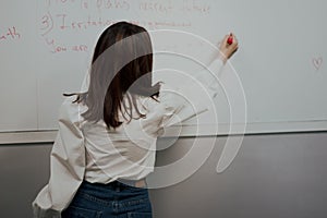 Back view of a cute young englisg language teacher in white blouse writing on a board and giving a class