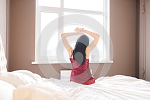 Back view. early morning caucasian brunette girl in red pajama stretching in bed after waking up