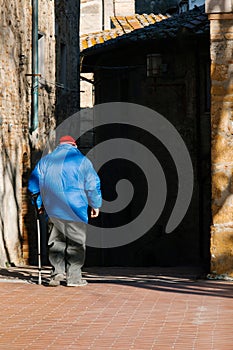 Back view of elderly stooped man with cane in the street of old city or town. Senior man in blue jacket and red hat with