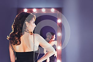 Back view elegant brunette in a black silk dressing standing in front of a tall make-up mirror in lilac color interior