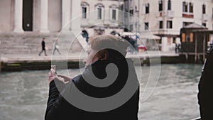 Back view excited active older Caucasian tourist woman taking smartphone photos on a gondola excursion on Venice trip.