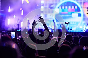 Back view of excited audience at night concert with hands raised, enjoying live music. Bright stage lights illuminate