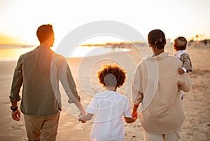Back view of family walking on the beach with two kids, holding hands, enjoying sunset on seaside