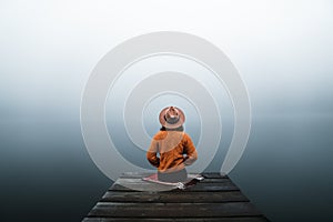 Back view of fashioned young woman sitting on wooden dock looking at view on a misty morning.