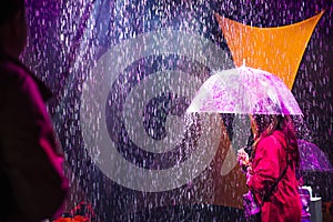 Back view female person with umbrella under artificial rain coming from the ceiling and illuminated by colored lights. Unusual